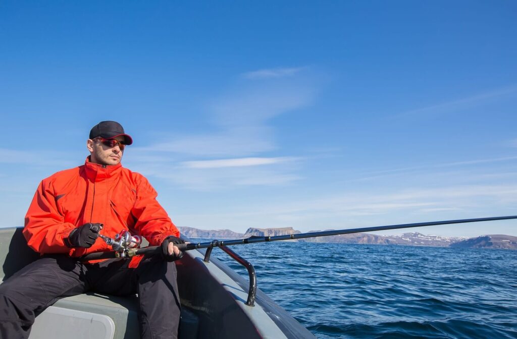 A fisherman wearing a red jacket casts their line over the edge of a boat. They are wearing polarized sunglasses to reduce glare.